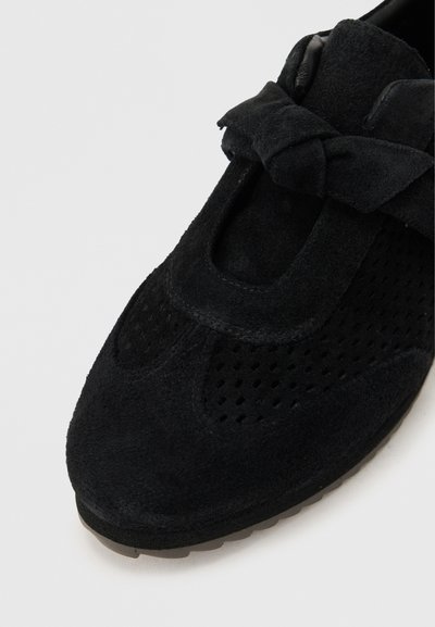 Μαύρο σουέτ παπούτσι με διάτρητες λεπτομέρειες και δεμένο φιόγκο πάνω στο λουρί, πάνω σε απλό φωτεινό φόντο.
