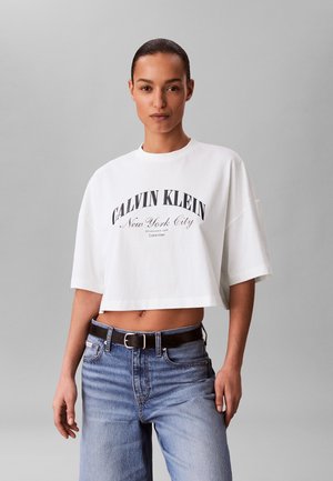 Femme aux cheveux tirés en arrière portant un t-shirt blanc raccourci Calvin Klein et un jean bleu avec ceinture noire, fond gris neutre.