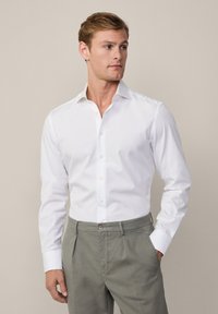 Weißes langärmliges Hemd mit spitzem Kragen und Knopfleiste, kombiniert mit hellgrauen Hosen. Baumwollstoff, taillierte Passform, glatte Textur.