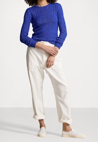 Kvinna i blå kabelstickad tröja, uppvikta vita byxor och vita sneakers, står med armarna korsade framför sig.