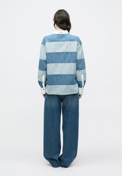 Chemise en jean avec des rayures horizontales bleu clair et bleu foncé, associée à un jean bleu foncé ample à jambes larges. Poignets blancs boutonnés visibles.