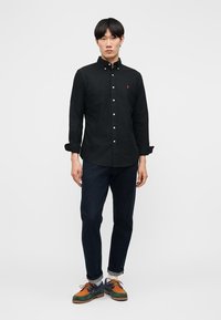 Modelo masculino con camisa negra abotonada, vaqueros oscuros remangados en los puños y zapatos con cordones multicolor en verde, naranja y azul.