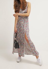 Květinové maxi šaty s bočním rozparkem, vyrobené z lehké látky v bílé, červené a zelené barvě. Nošeny s bílými teniskami a držena černá taška.