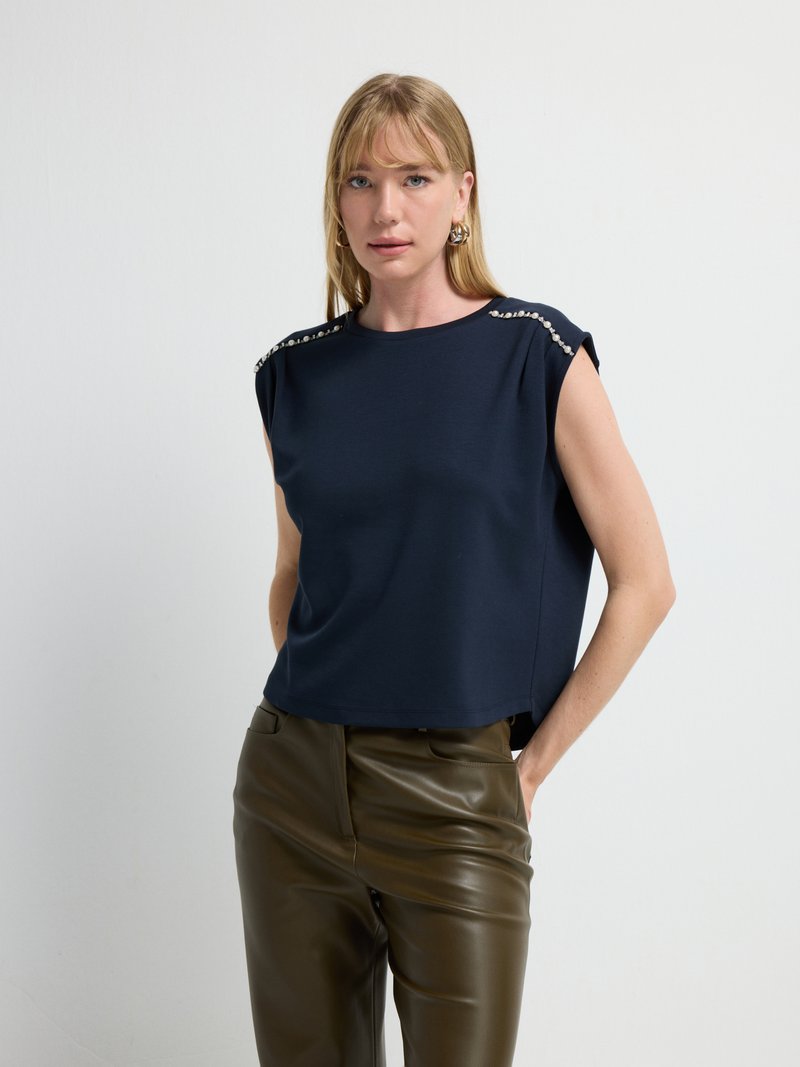 Top senza maniche blu navy con una vestibilità rilassata, decorato con accessori sui lati e realizzato in tessuto morbido e liscio. Indossato con pantaloni marroni lucidi.