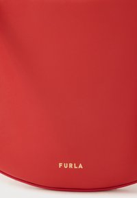 Κόκκινη δερμάτινη τσάντα με λεία υφή, που διαθέτει στρογγυλεμένο σχήμα και διακριτικό λογότυπο της μάρκας σε χρυσό στο κάτω μέρος.