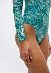 Κοντινό πλάνο ενός ατόμου που φοράει μία πετρόλ μακρυμάνικη φόρμα κολύμβησης με ανοιχτόχρωμο τροπικό σχέδιο φύλλων και εμφανές λογότυπο του εμπορικού σήματος στον καρπό.