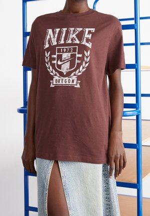 Person iført en brun Nike t-shirt med teksten "1972 Oregon", kombineret med et lyst denimskørt med en frontslids, stående ved blå hylder.