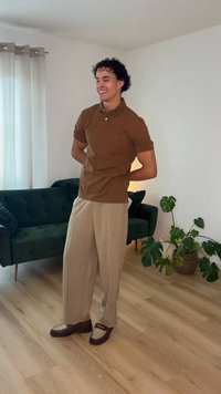 Νέος άνδρας που χαμογελά με τα χέρια πίσω από την πλάτη, φοράει καφέ πόλο, μπεζ παντελόνι και μοκασίνια, στέκεται σε ένα φωτεινό σαλόνι με πράσινο καναπέ και φυτά.