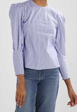 Donna che indossa una blusa a righe verticali bianche e blu con maniche lunghe a sbuffo e jeans blu, in piedi contro uno sfondo semplice.