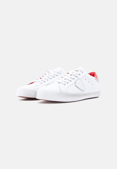 Λευκά αθλητικά παπούτσια με λείο επάνω μέρος, στρογγυλή μύτη, αντίθετες κόκκινες λεπτομέρειες στο τακούνι και διακριτική λεπτομέρεια με λογότυπο στο πλάι.