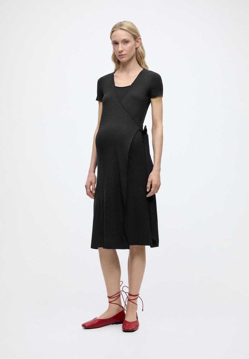 Εγκυος γυναίκα με μαύρο φόρεμα τύπου wrap με κοντά μανίκια και κόκκινα παπούτσια με κορδόνια, στέκεται μπροστά σε λευκό φόντο.