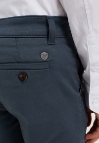 Námořnické modré kalhoty vyrobené z hladké látky, s kovovým stříbrným logem, hnědým knoflíkovým zapínáním a skrytou boční kapsou.