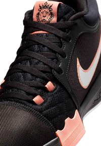 Chaussure de sport noire et rose avec le swoosh Nike blanc, des lacets noirs et un logo tête de lion sur l'étiquette rose de la languette.