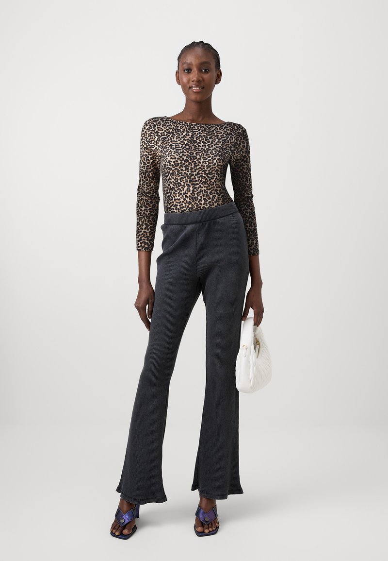 Langärmliges Oberteil mit Leopardenmuster, schwarze gerippte Hose und lila High Heels. Das Model hält eine weiße, gesteppten Handtasche und steht vor einem neutralen Hintergrund.