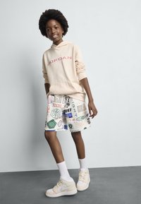 Haut à capuche beige clair avec "JORDAN" en rouge, short multicolore à motifs, chaussettes blanches mi-mollet et baskets montantes beiges.