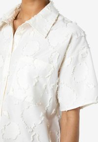 Camicia bianca con un motivo testurizzato e dettagli sfrangiati, maniche corte e una tasca sul petto. Realizzata in tessuto leggero.