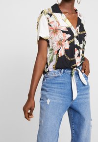 Kortärmad blus i svart med blommönster i rosa och grönt, med krage och knappstängning, knuten vid midjan.