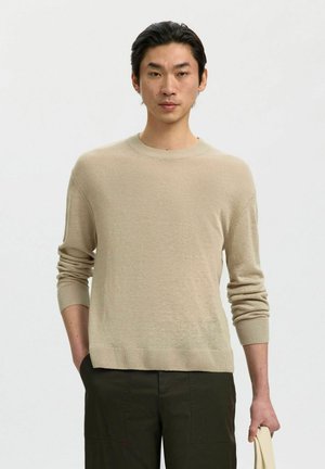 Junger Mann mit dunklen Haaren, der einen beigen Langarmpullover und dunkle Hosen trägt, hält eine hellfarbige Tragetasche in der rechten Hand.