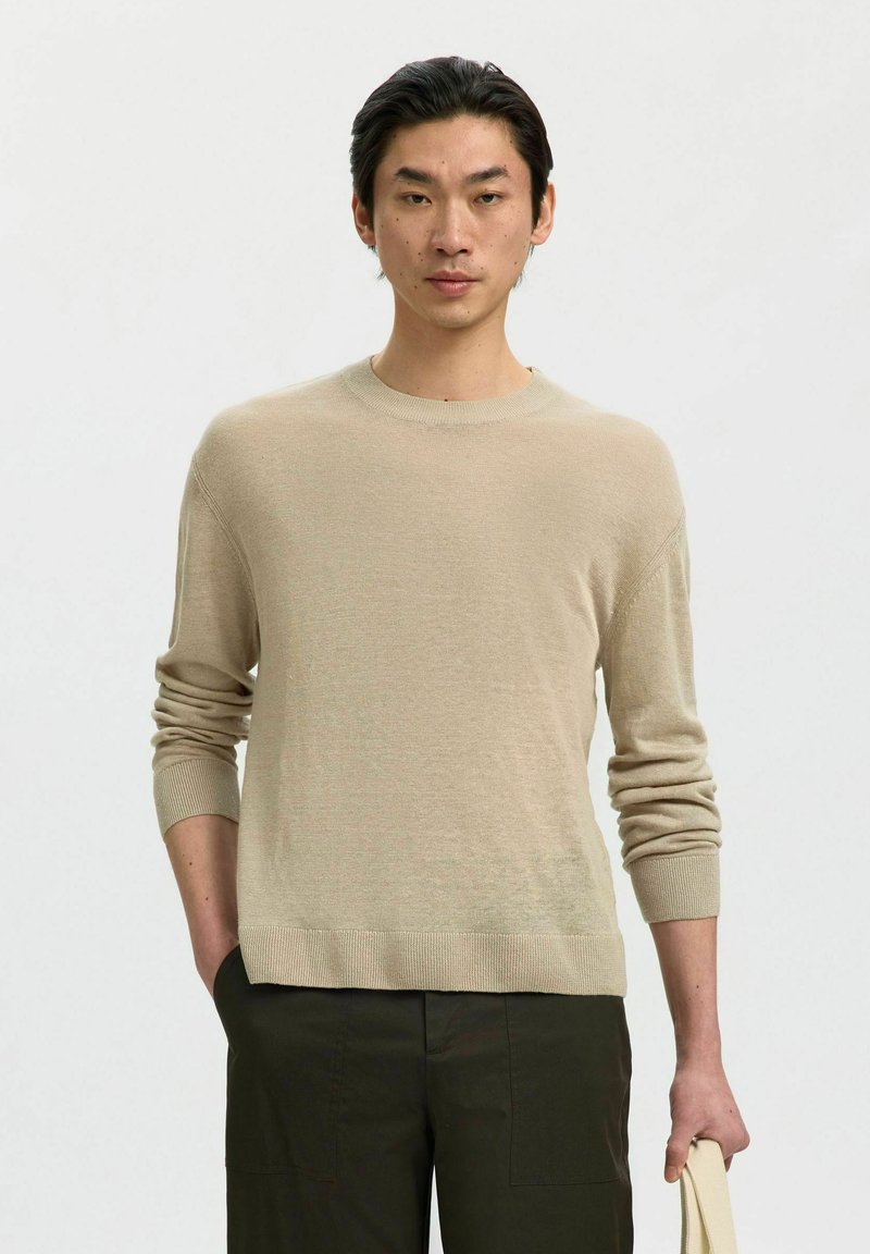Jeune homme aux cheveux foncés portant un pull beige à manches longues et un pantalon foncé, tenant un sac fourre-tout clair dans sa main droite.
