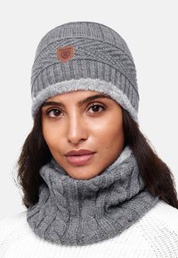 Bonnet en tricot gris avec une bande en fausse fourrure et un patch logo en cuir. Associé à un col roulé texturé gris assorti. Matériaux doux et confortables.