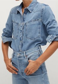Chemise en denim bleu clair avec deux poches poitrine, boutons sur le devant, manches retroussées, associée à un jean en denim bleu taille haute.