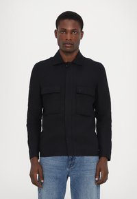 Marineblaue Knopfjacke mit zwei Brusttaschen, kragenlosem Hals, langen Ärmeln und geradem Schnitt, aus einem glatten Stoff gefertigt.