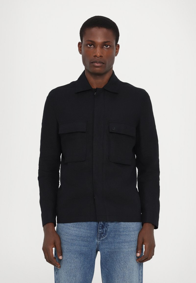 Marineblaue Knopfjacke mit zwei Brusttaschen, kragenlosem Hals, langen Ärmeln und geradem Schnitt, aus einem glatten Stoff gefertigt.