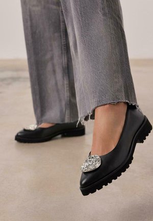 Chaussures en cuir noir avec un bout pointu, ornées d'un embellissement décoratif en argent à l'avant et d'une semelle en caoutchouc texturée.