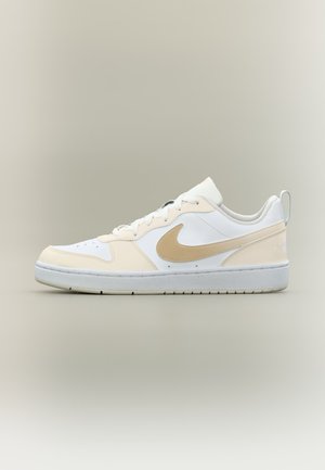 Zemu profilu balti sporta apavi ar bēša krāsas pārklājumiem, bēša Nike svītru, baltām auklām un baltu zolīti uz neitrāla fona.