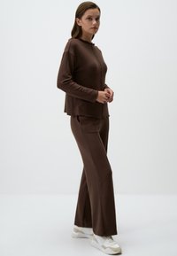 Braunes Strickoutfit mit einem lockeren Oberteil mit Rundkragen und langen Ärmeln, kombiniert mit weit geschnittenen Hosen. Weiße Sneakers mit beigen Akzenten.