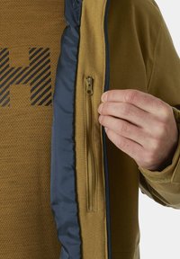 Giacca marrone con zip e fodera blu, caratterizzata da uno strato interno testurizzato e una mano che regola la zip sul lato sinistro.