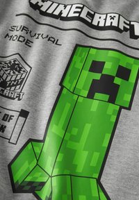 Grijze katoenen t-shirt met een grote groene pixelachtige Creeper-figuur uit Minecraft, met zwarte en groene tekst die "Survival Mode" vermeldt.