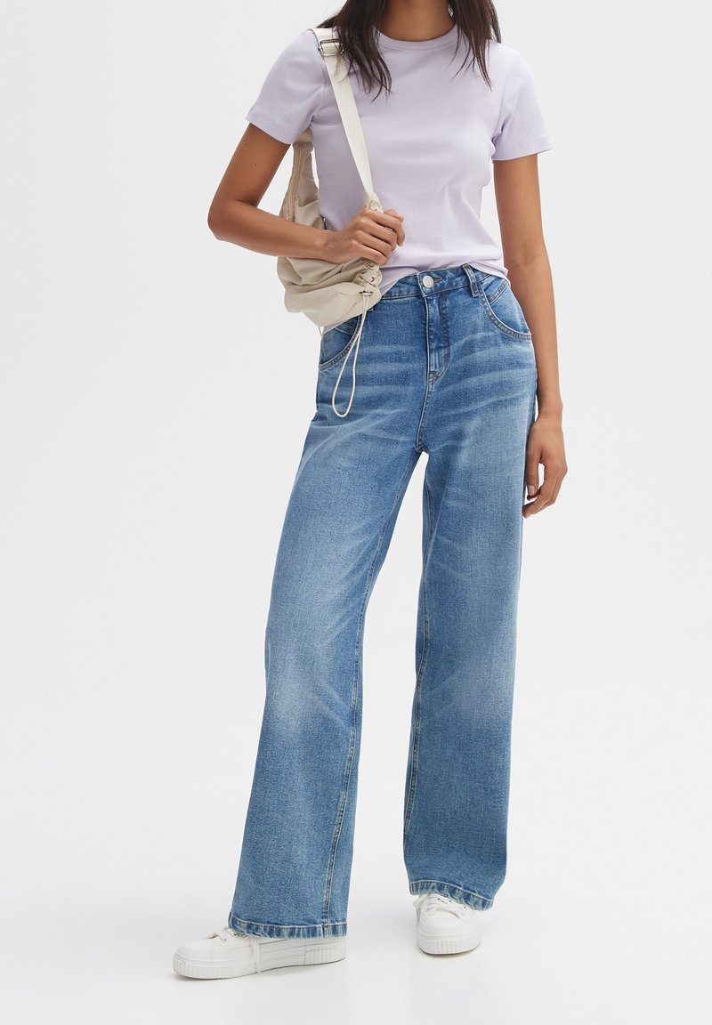 Wide-leg blue denim jeans, high-waisted with light fading, paired with a lavender short-sleeve shirt and white sneakers. Linked beige bag.