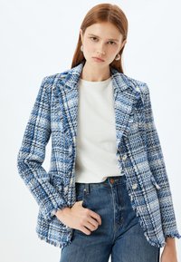 Vävd blå och vit kavaj med fransad nederkant, dubbla knappar och texturerat mönster. Kombinerad med en vit topp och blå jeans.