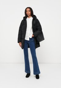 Schwarze gefütterte Jacke mit Kapuze, featuring ein quiltetes Muster. Getragen über einem weißen Hemd mit Knopfleiste und kombiniert mit weit geschnittenen blauen Jeans.