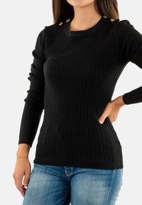 Donna che indossa un maglione aderente nero a trecce con bottoni dorati sulle spalle e jeans blu, in piedi con una mano sul fianco.