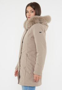 Beige winterjas met een faux fur capuchon, ritssluiting aan de voorkant, zijzakken en een zachte, gladde textuur. Middenlange ontwerp geschikt voor koud weer.