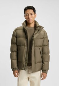 Olivengrønt puffer-jakke med høj krave, lynlås og trykknaplukning, quiltet design og ribbede manchetter. Båret over en struktureret brun sweater.
