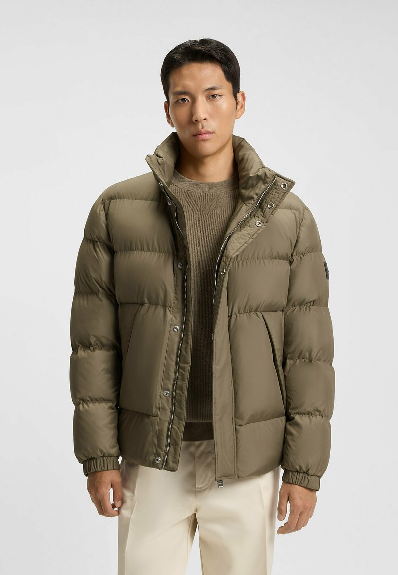 Olivengrønt puffer-jakke med høj krave, lynlås og trykknaplukning, quiltet design og ribbede manchetter. Båret over en struktureret brun sweater.
