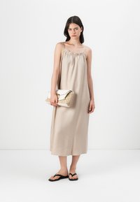 Bež maxi haljina s naramenicama i naboranim dekolteom. Uparena s kremastom torbom s teksturom u zlatnoj boji i crnim japankama.