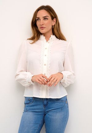 Blusa bianca realizzata in tessuto leggero con colletto arricciato, chiusura con bottoni e rifinitura in pizzo. Maniche lunghe con dettagli in pizzo nei polsini.