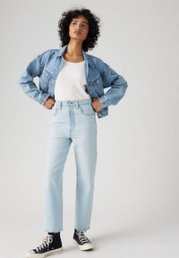 Άτομο στέκεται με τα χέρια στη μέση φορώντας ανοιχτό μπλε τζιν μπουφάν, ψηλόμεσα ανοιχτό μπλε τζιν παντελόνια, λευκό πουκάμισο και μαύρα αθλητικά παπούτσια.