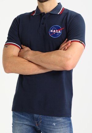 Uomo che indossa una polo NASA blu navy con bordi rossi e bianchi, con le braccia incrociate, abbinata a jeans blu.