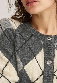 Harmaa ja kerma valkoinen argyle-kuviollinen neuletakki, joka on valmistettu teksturoidusta villasta. Siinä on pyöreä pääntie ja kaksi harmaata nappia edessä.