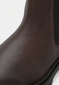 Cizme din piele maro, până la gleznă, cu o suprafață texturată, panouri laterale elastice și vârf rotund. Prezintă o talpă din cauciuc negru.