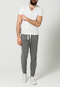 Camiseta blanca de cuello en V con pantalones deportivos grises que tienen bolsillos laterales y cordón blanco. Modelo con zapatillas blancas; fondo liso.