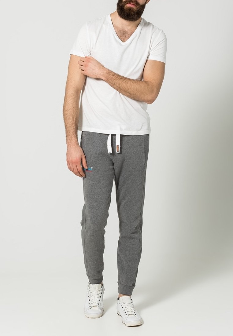 Camiseta blanca de cuello en V con pantalones deportivos grises que tienen bolsillos laterales y cordón blanco. Modelo con zapatillas blancas; fondo liso.