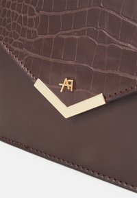 Brun väska i konstläder med ett texturerat krokodilmönster, som har en guldfärgad metallaccent i form av en chevron och sydda kanter.