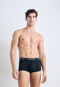 Oberkörperfreier Mann mit lockigem Haar, der schwarze Calvin Klein Boxer-Shorts trägt und vor einem schlichten hellgrauen Hintergrund steht.