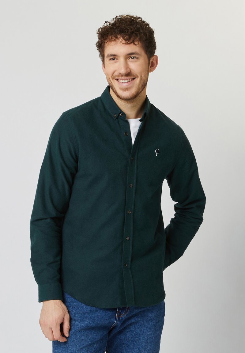 Donkergroene button-up overhemd met lange mouwen, voorzien van een klein wit logo op de borst en een getailleerde pasvorm, gecombineerd met blauwe spijkerbroeken.